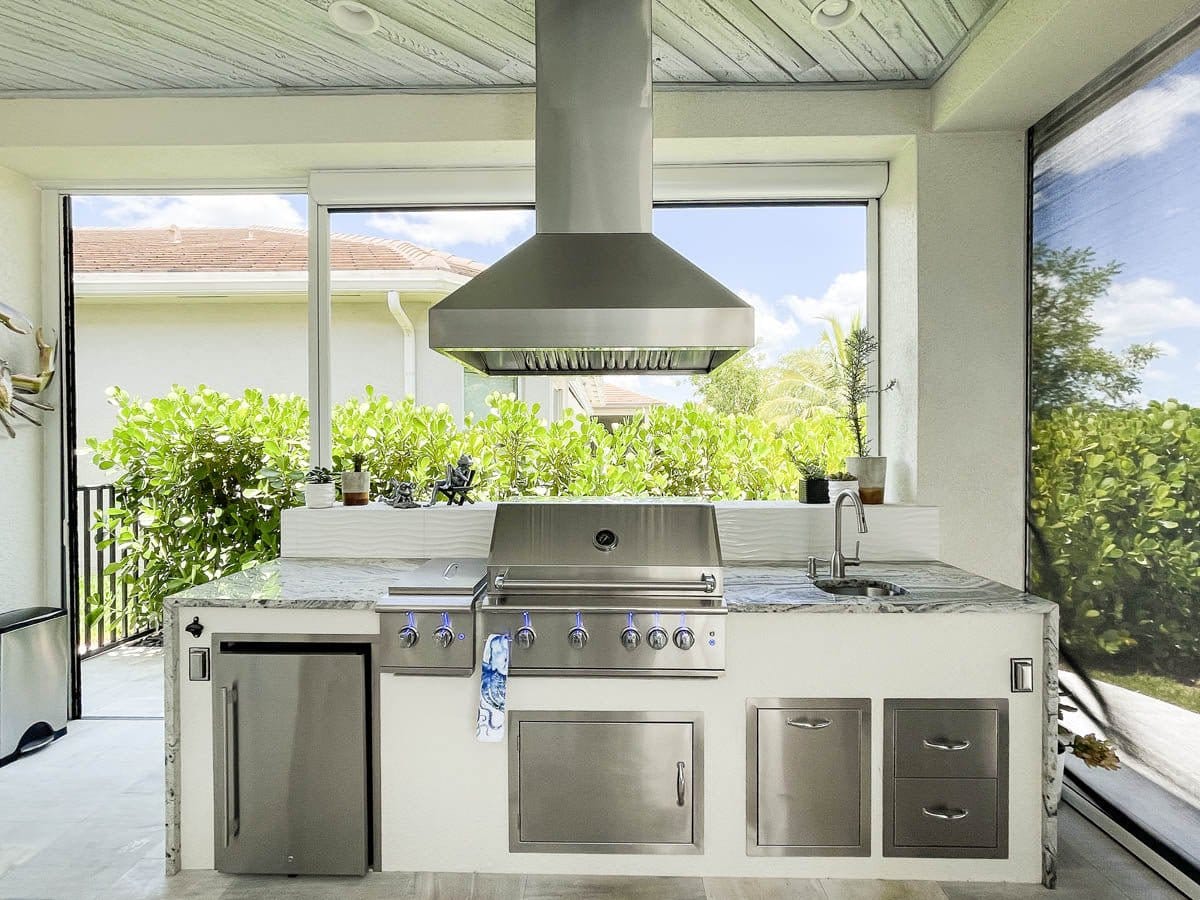 An outdoor kitchen featuring ProlineRangeHoods.com PLJI 103 Island Hood with 1200 CFM, robust cabinets, and verdant greenery.
