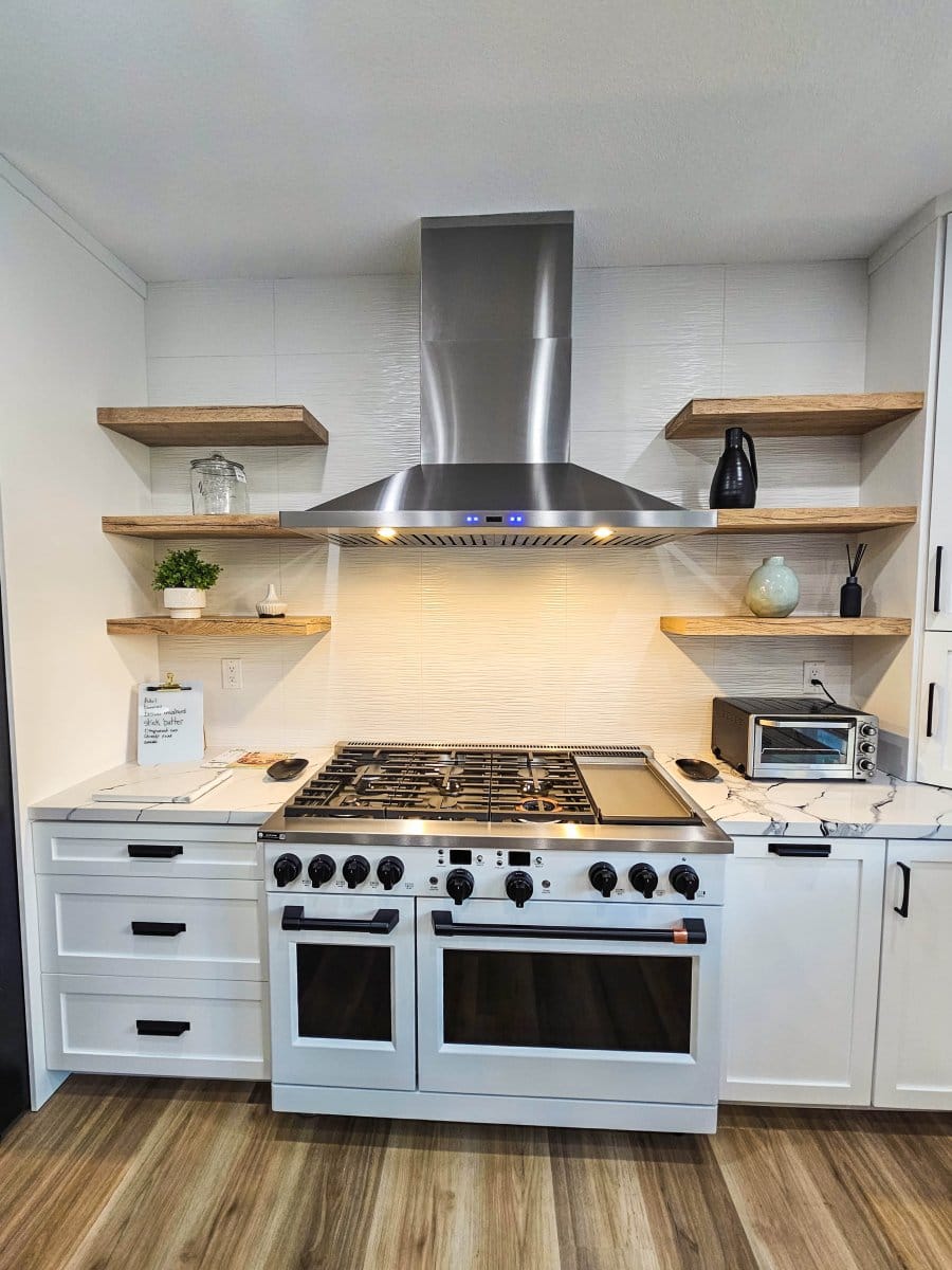 A contemporary kitchen with ProlineRangeHoods.com stainless steel stove, wooden shelves, and white cabinets on a wooden floor.