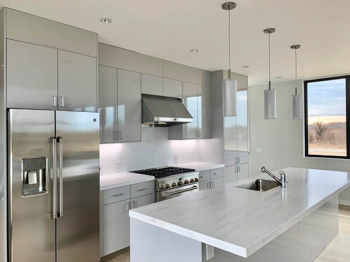 A modern kitchen featuring gray cabinets, ProlineRangeHoods.com's sleek under-cabinet range hood (PLFW 832), an island sink, and pendant lights.