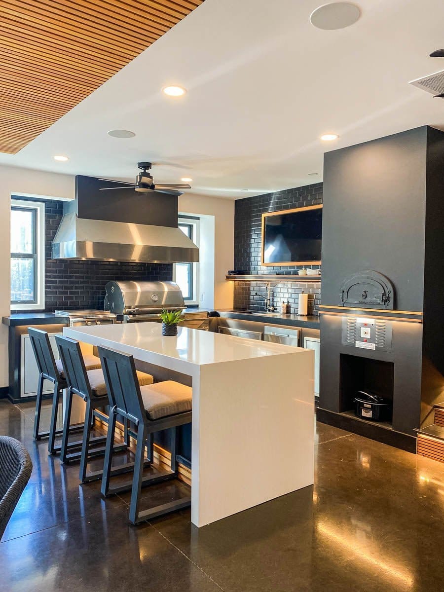 Step into a modern kitchen with ProlineRangeHoods.com, stylish bar stools, stainless steel appliances, and a wall-mounted TV.