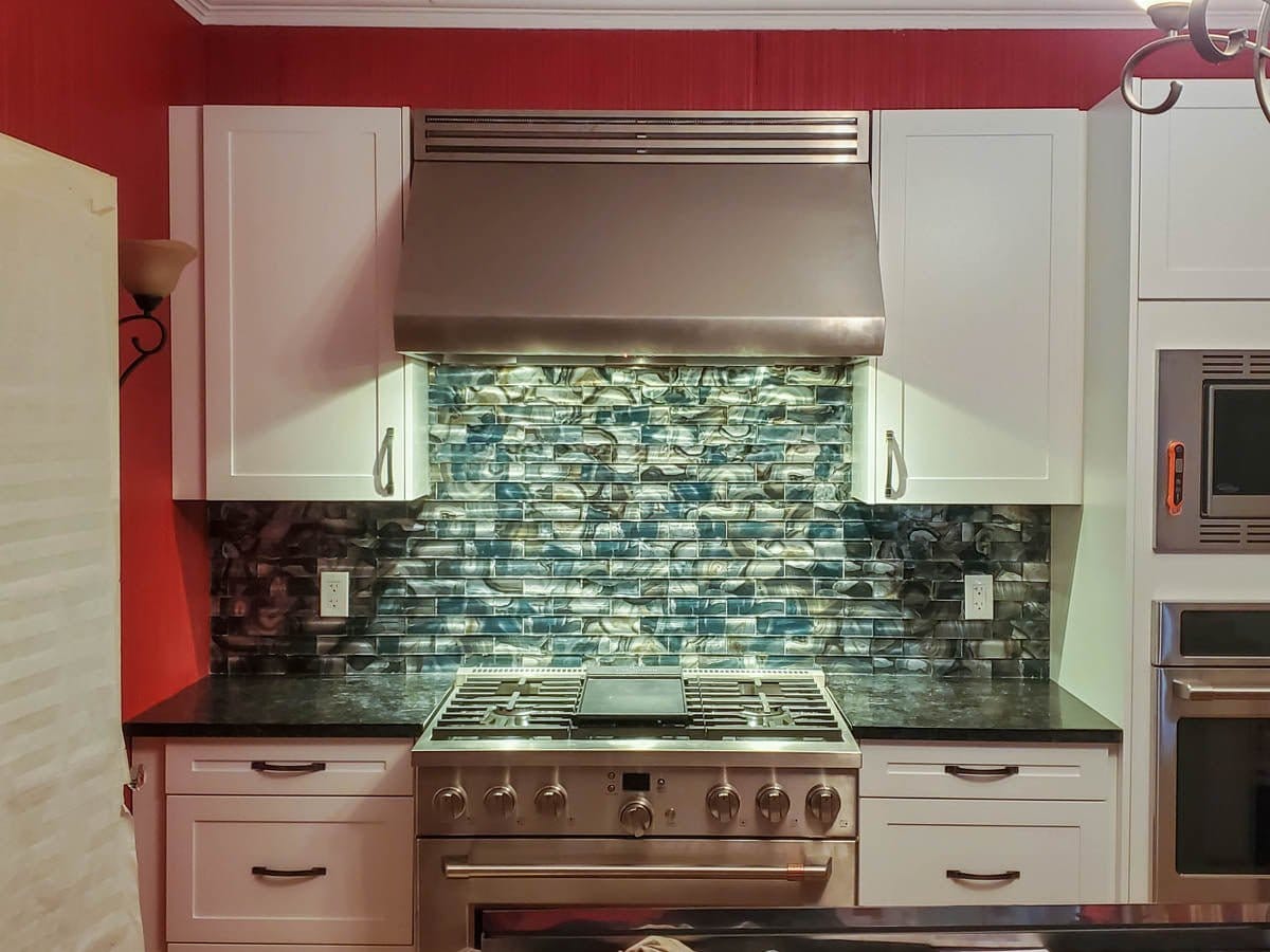 Kitchen with ProlineRangeHoods.com PLJW 109 hood, white cabinets, dark counters, and teal glass tile backsplash.