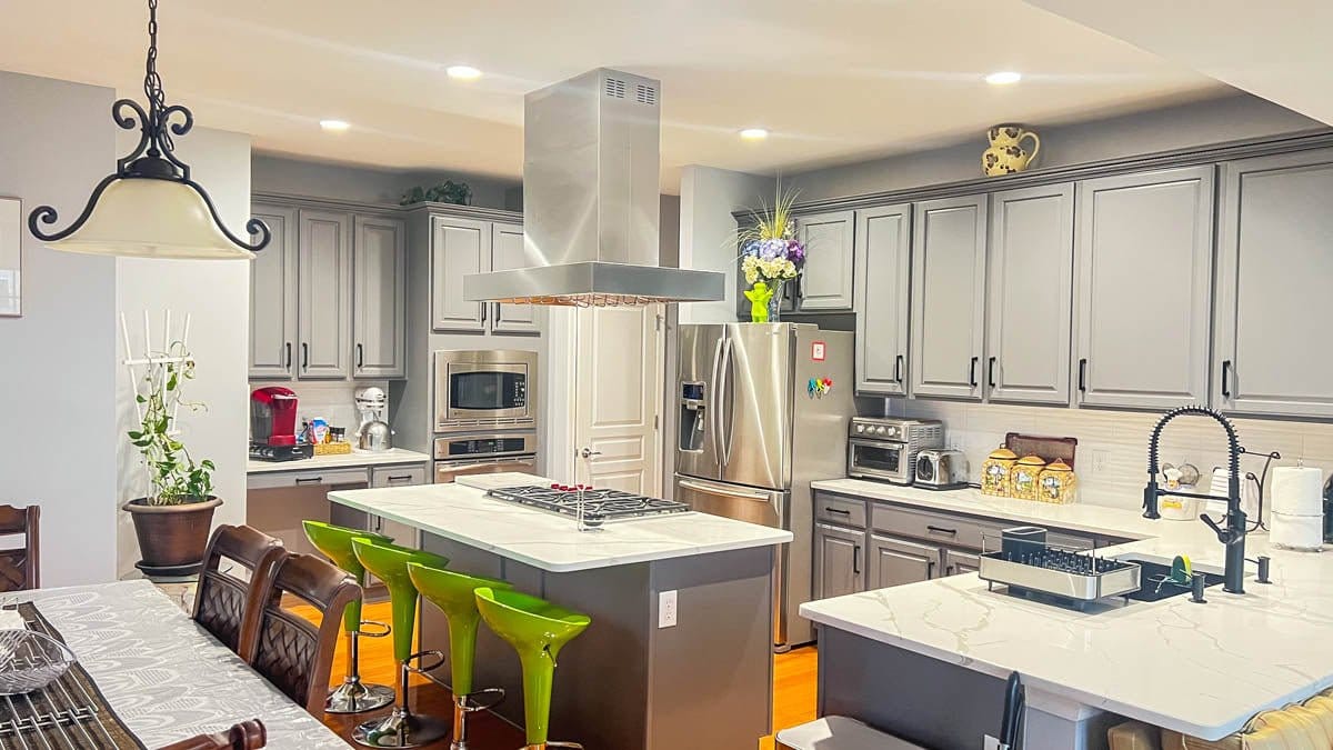 The modern kitchen features gray cabinets, stainless steel appliances, and green barstools. ProlineRangeHoods.com.
