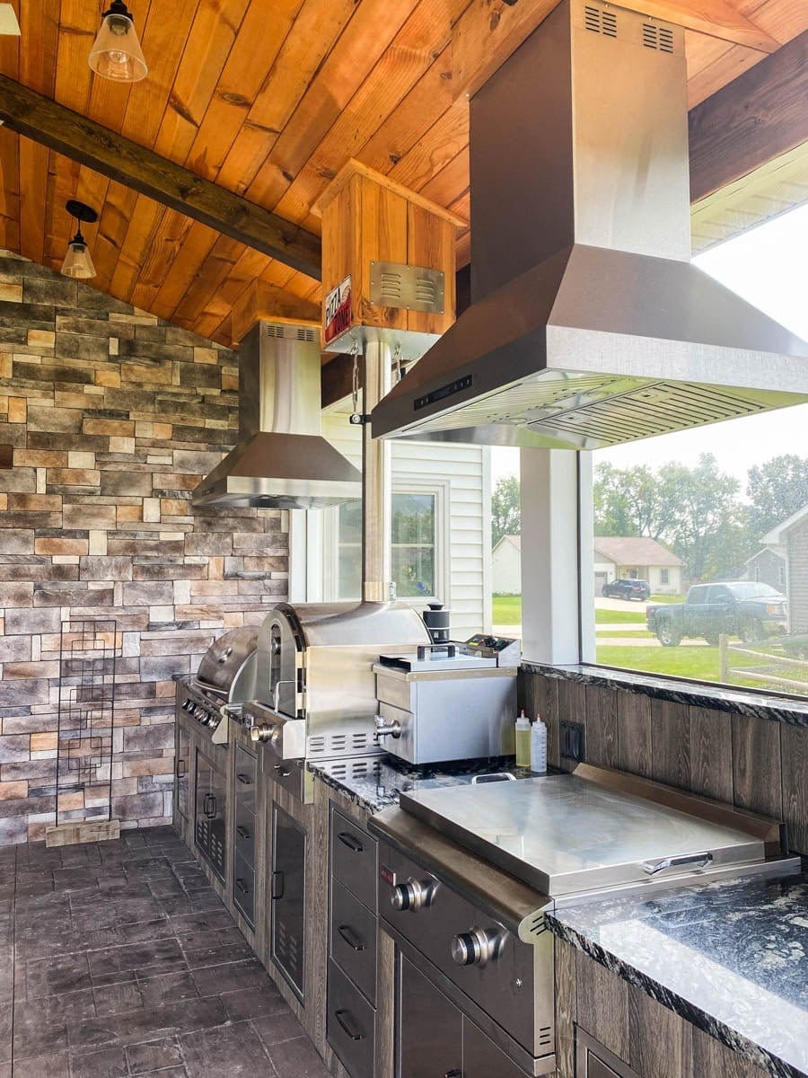 Outdoor kitchen with ProlineRangeHoods.com Island Range Hood, stainless finish, LCD controls, stone wall, wood ceiling.
