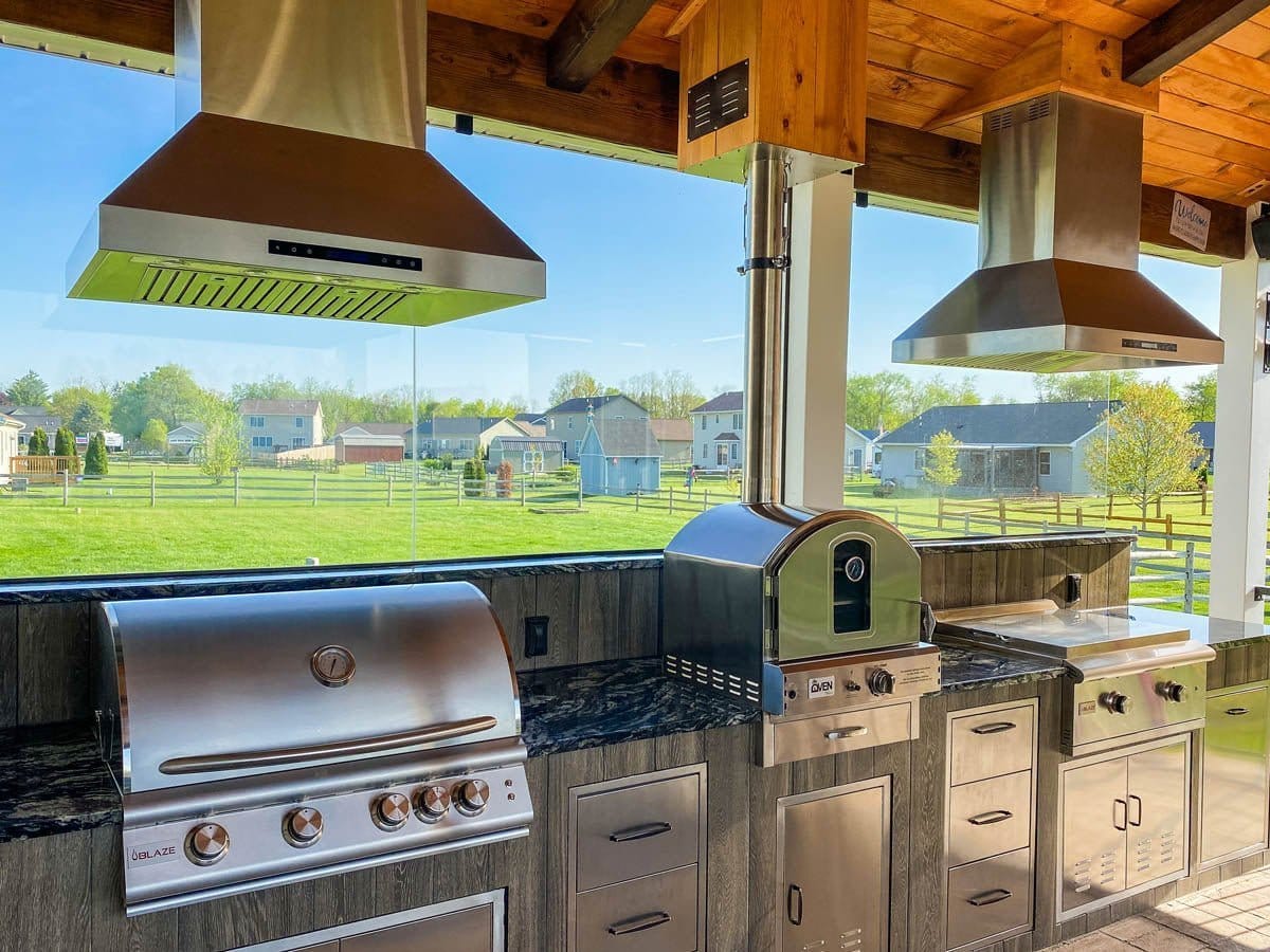An outdoor kitchen with ProlineRangeHoods.com showcases the Proline PROSI Island Range Hood under a wooden roof.
