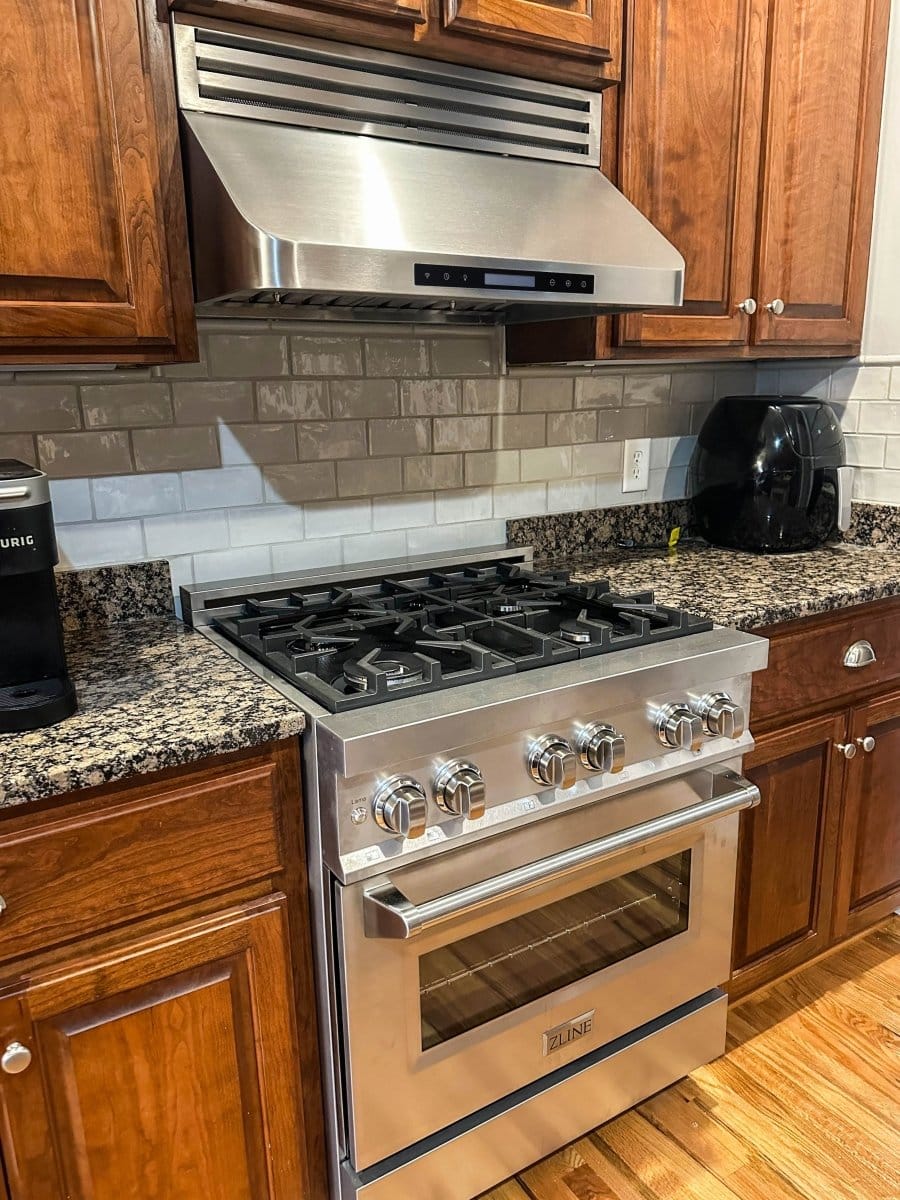 Modern kitchen with a stainless stove, ProlineRangeHoods.com PLFW 115 range hood, wood cabinets, granite countertops.