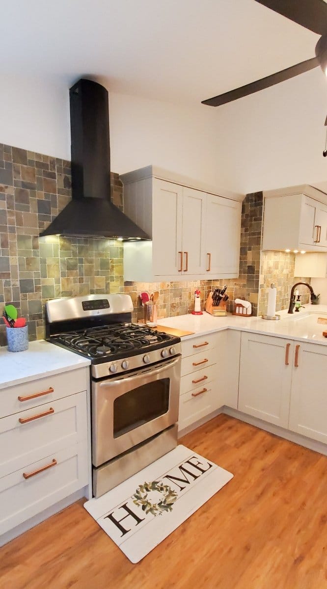 A modern kitchen with a Proline Vector hood from ProlineRangeHoods.com, tile backsplash, white cabinets, and a "HOME" mat.