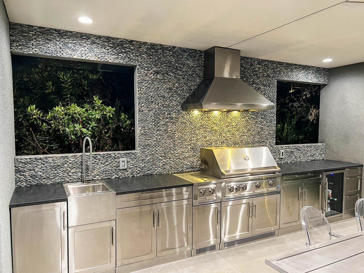 Modern outdoor kitchen at night with stainless appliances, grill, Proline PLJW 109 WC hood, stone backsplash. ProlineRangeHoods.com.